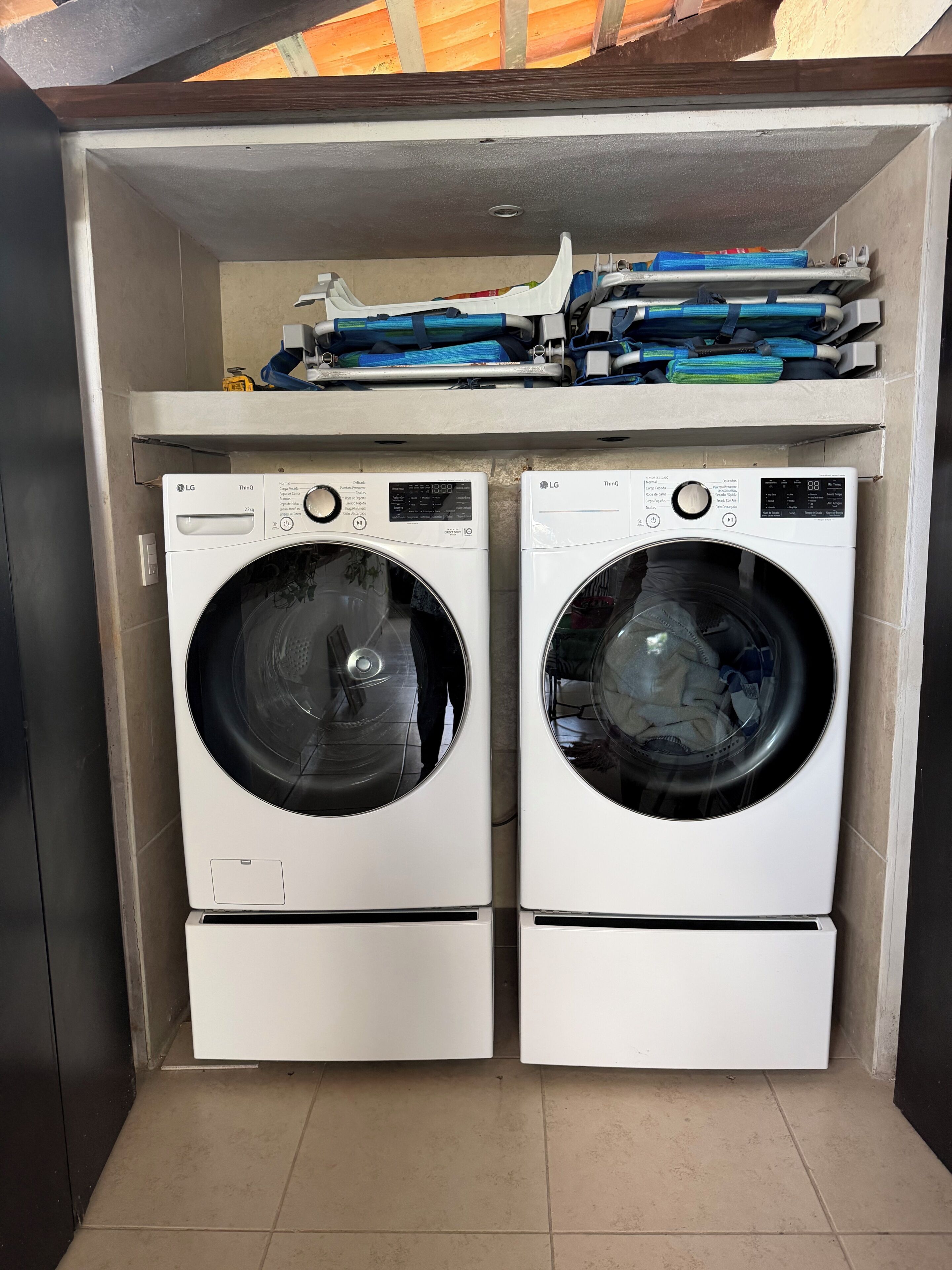 A laundry room with two white LG washing machines, a shelf with folded clothes, and a tiled floor.