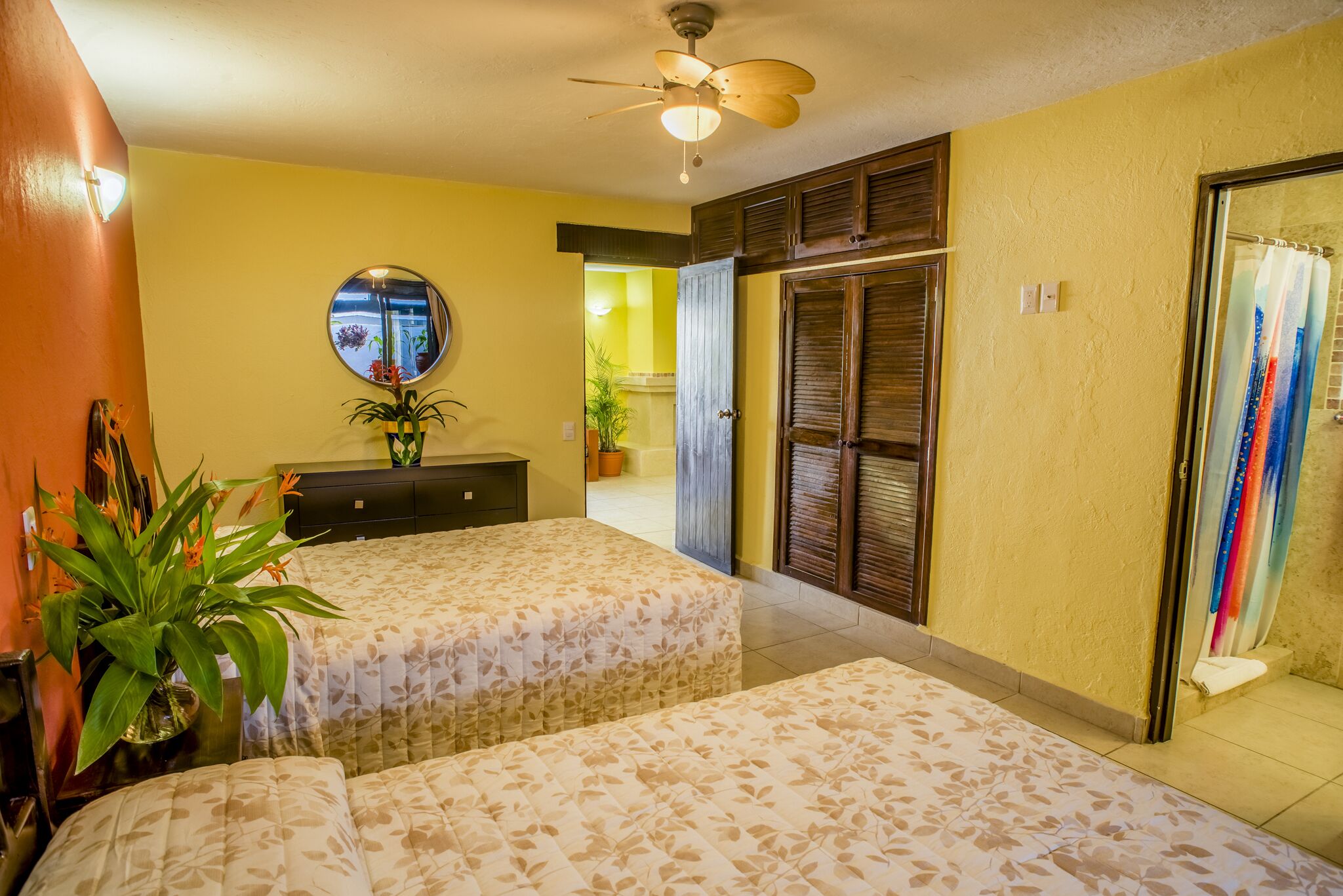 A hotel room with two beds, a ceiling fan, a mirror, a potted plant, and a sliding door leading to a bathroom.
