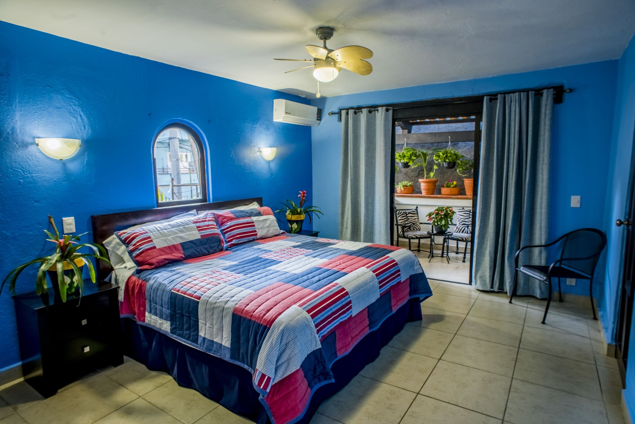 Une chambre avec un mur bleu, un lit recouvert d’une couette à rayures, un ventilateur de plafond et une vue sur un balcon agrémenté de plantes.