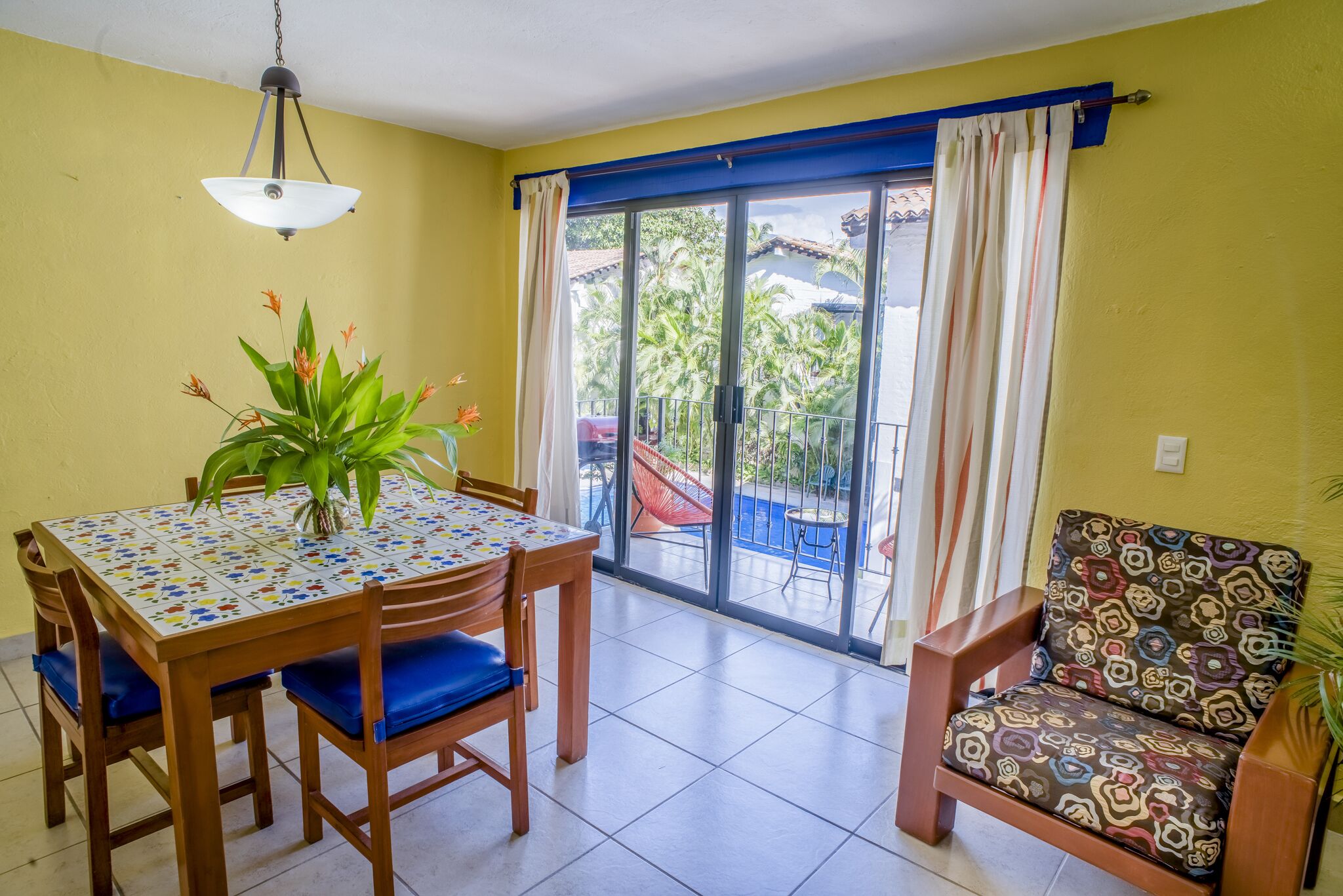 A dining area with a table and chairs, a hanging light fixture, and a view through a sliding glass door to an outdoor area with chairs and a pool.