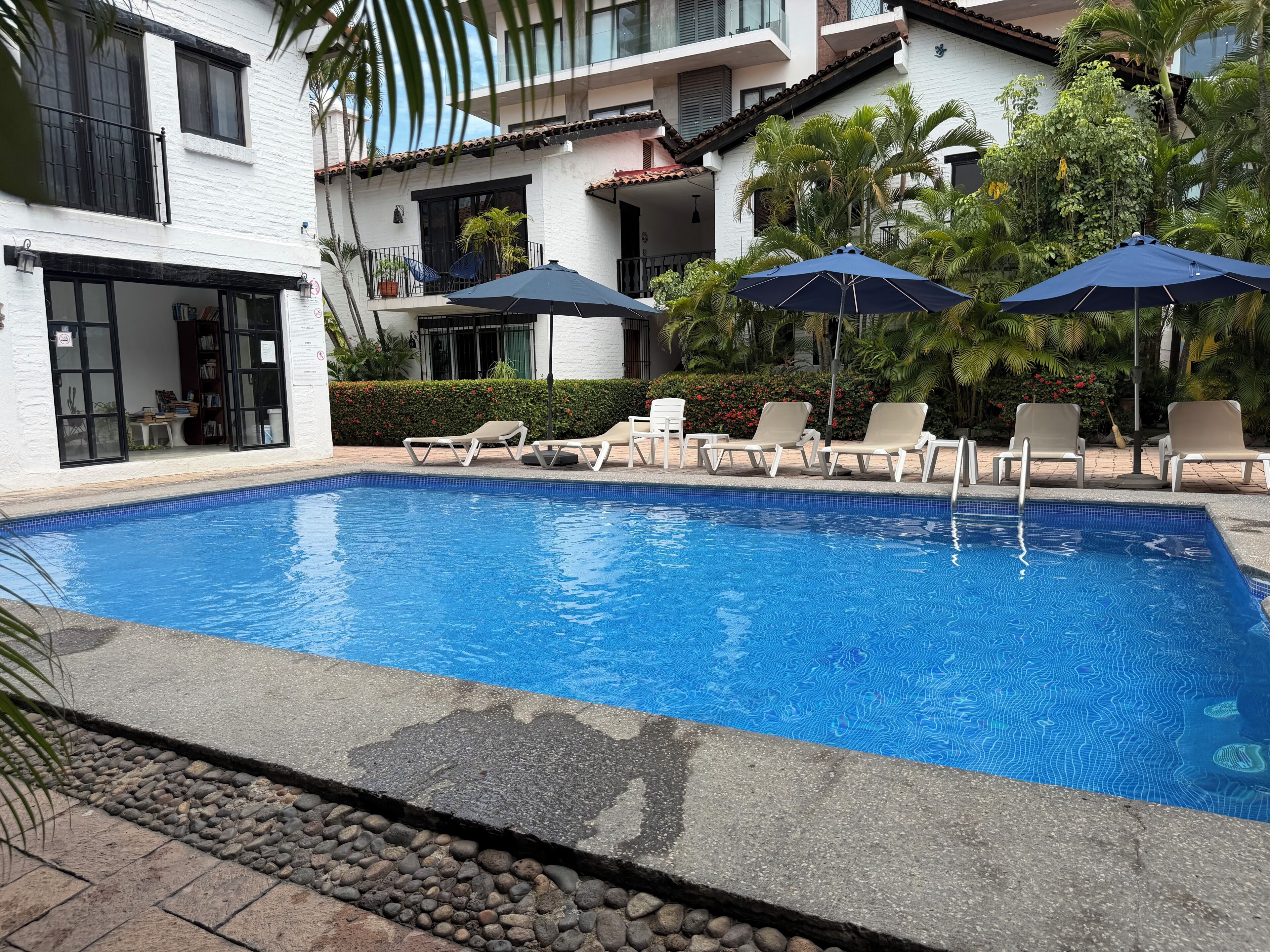 A swimming pool with lounge chairs and umbrellas, surrounded by a building with balconies and greenery.