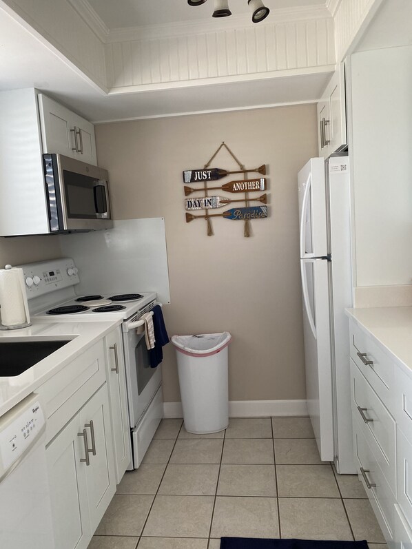 A compact kitchen with white cabinetry, a microwave, stove, and a refrigerator. There is a trash bin and a wall decoration with wooden paddles.