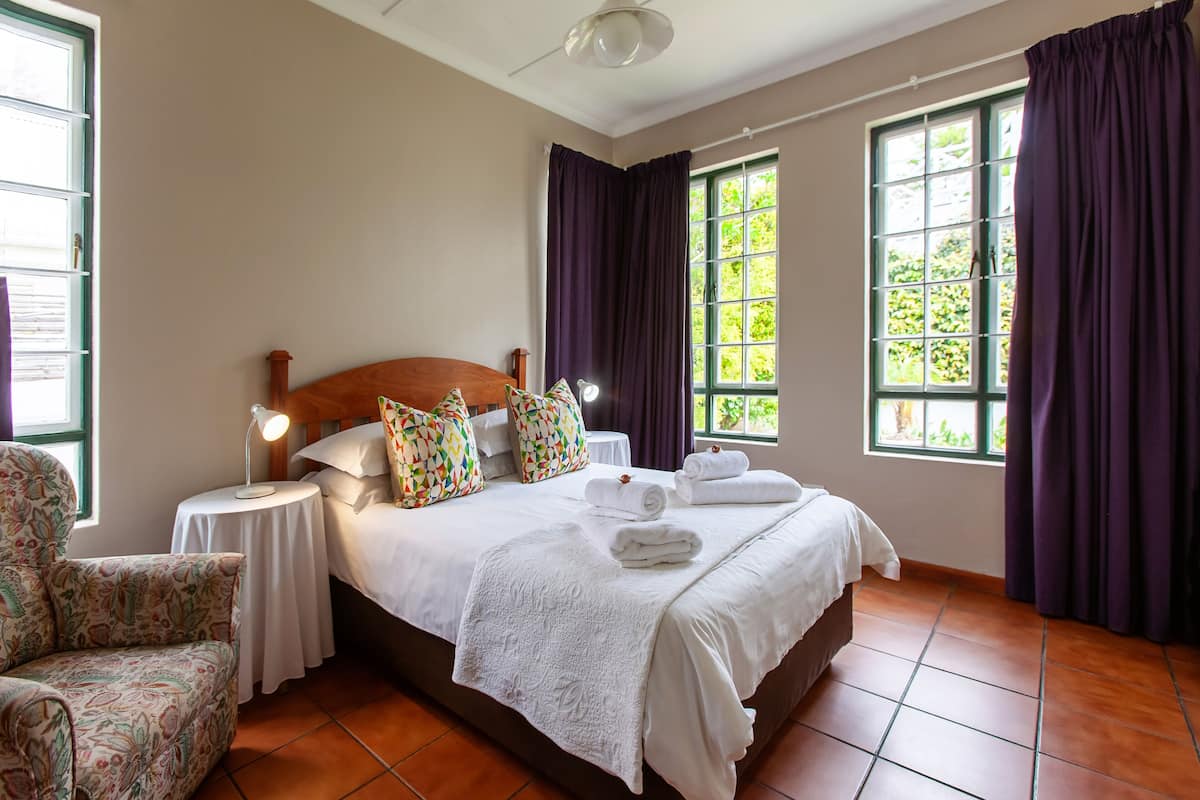 A neatly arranged hotel room with a large bed, a bedside table with a lamp, a chair, and two windows with curtains.