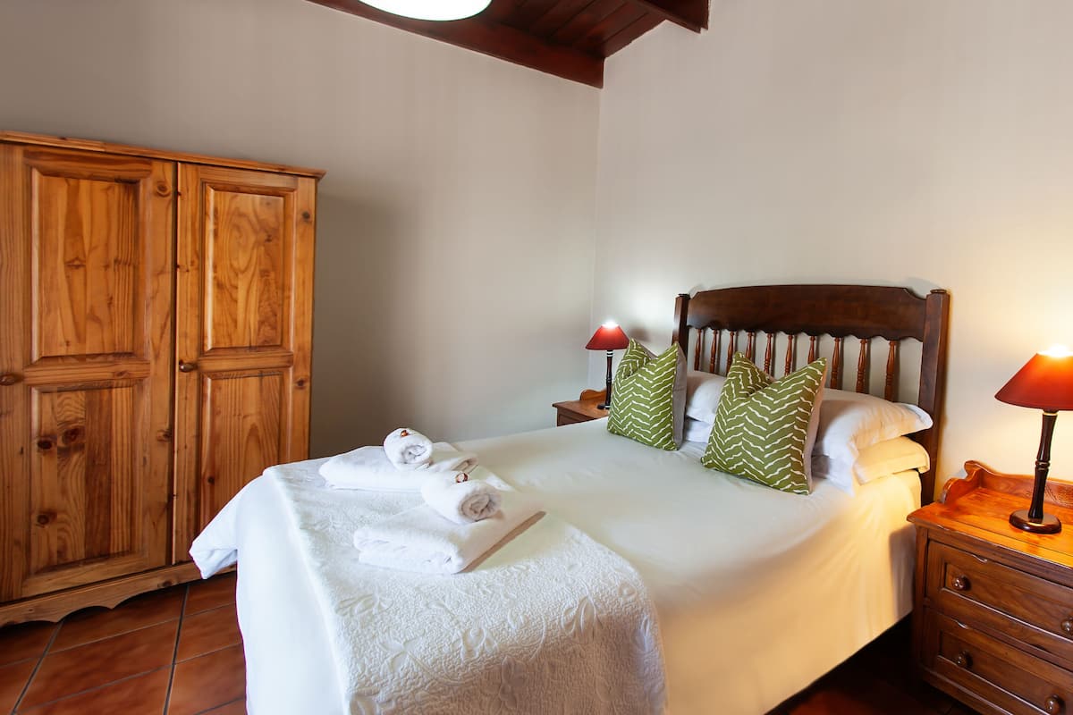 A neatly made bed with white linens and rolled towels, a wooden wardrobe, and a nightstand with a lamp.