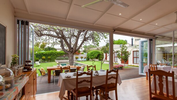 A dining area with tables and chairs, a view of a garden with a tree, and a pool in the background. - The Habit Self Catering (Worcester)