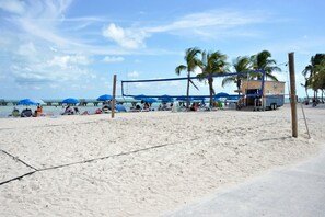 A beach volleyball court with a net, palm trees, and beach umbrellas.