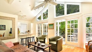 A spacious living area with a high ceiling, a ceiling fan, a dining table, two green armchairs, a brown leather sofa, and large windows with a view of greenery outside. - Key West Dreamin` - Private Pool Steps Off Duval (Key West)