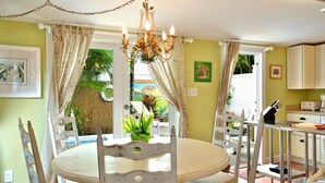 A dining area with a round table, white chairs, a chandelier, and a view of a tropical garden through the window. - Key West Wabi Sabi - Authentic Downtown Cottage w Pool - Steps Off Duval (Key West)