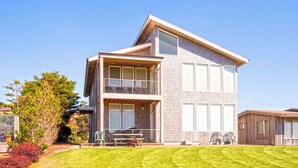 A two-story modern house with a balcony on the upper floor, a well-maintained lawn, and a clear sky. - OCEAN FRONT: MODERN MARVEL: Seaside Vacation Homes, Inc Managed Property (Gearhart)