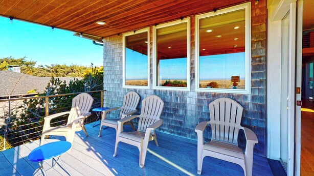 A covered outdoor area with Adirondack chairs and a blue table, overlooking a scenic landscape. - OCEAN FRONT: MODERN MARVEL: Seaside Vacation Homes, Inc Managed Property (Gearhart)