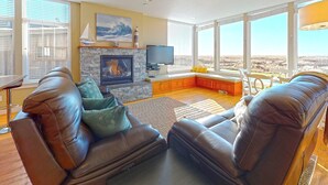 A living room with leather sofas, a stone fireplace, a television, and a view of the landscape through large windows. - OCEAN FRONT: MODERN MARVEL: Seaside Vacation Homes, Inc Managed Property (Gearhart)