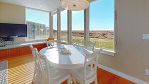A dining area with a white table and chairs, a television, and large windows with a view of a field. - OCEAN FRONT: MODERN MARVEL: Seaside Vacation Homes, Inc Managed Property (Gearhart)