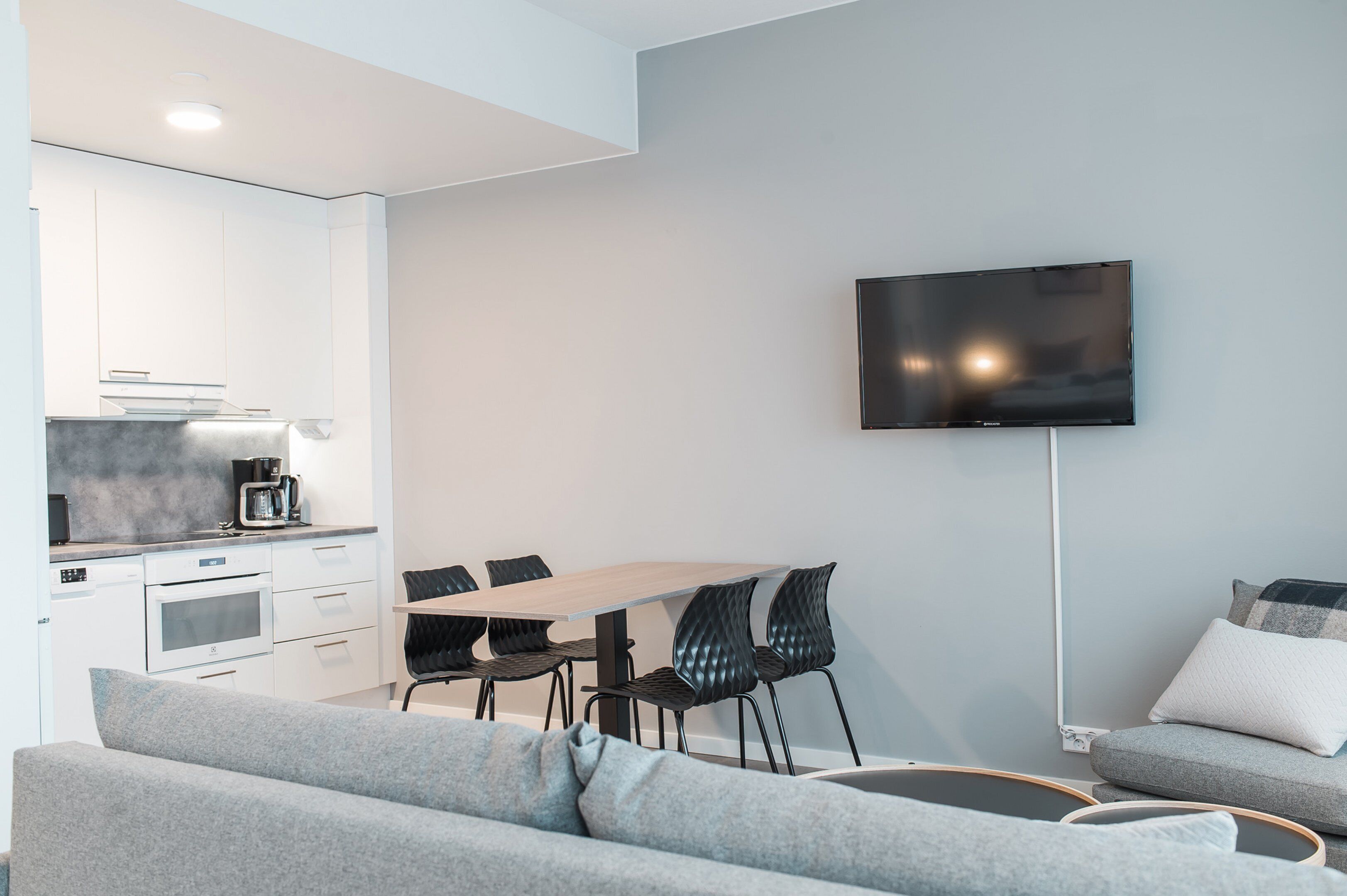 Una sala moderna con un sofá gris, una mesa de comedor con sillas, un televisor de pantalla plana montado en la pared y una cocina con armarios blancos y una cafetera.