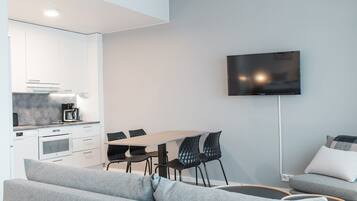 Una sala moderna con un sofá gris, una mesa de comedor con sillas, un televisor de pantalla plana montado en la pared y una cocina con armarios blancos y una cafetera.