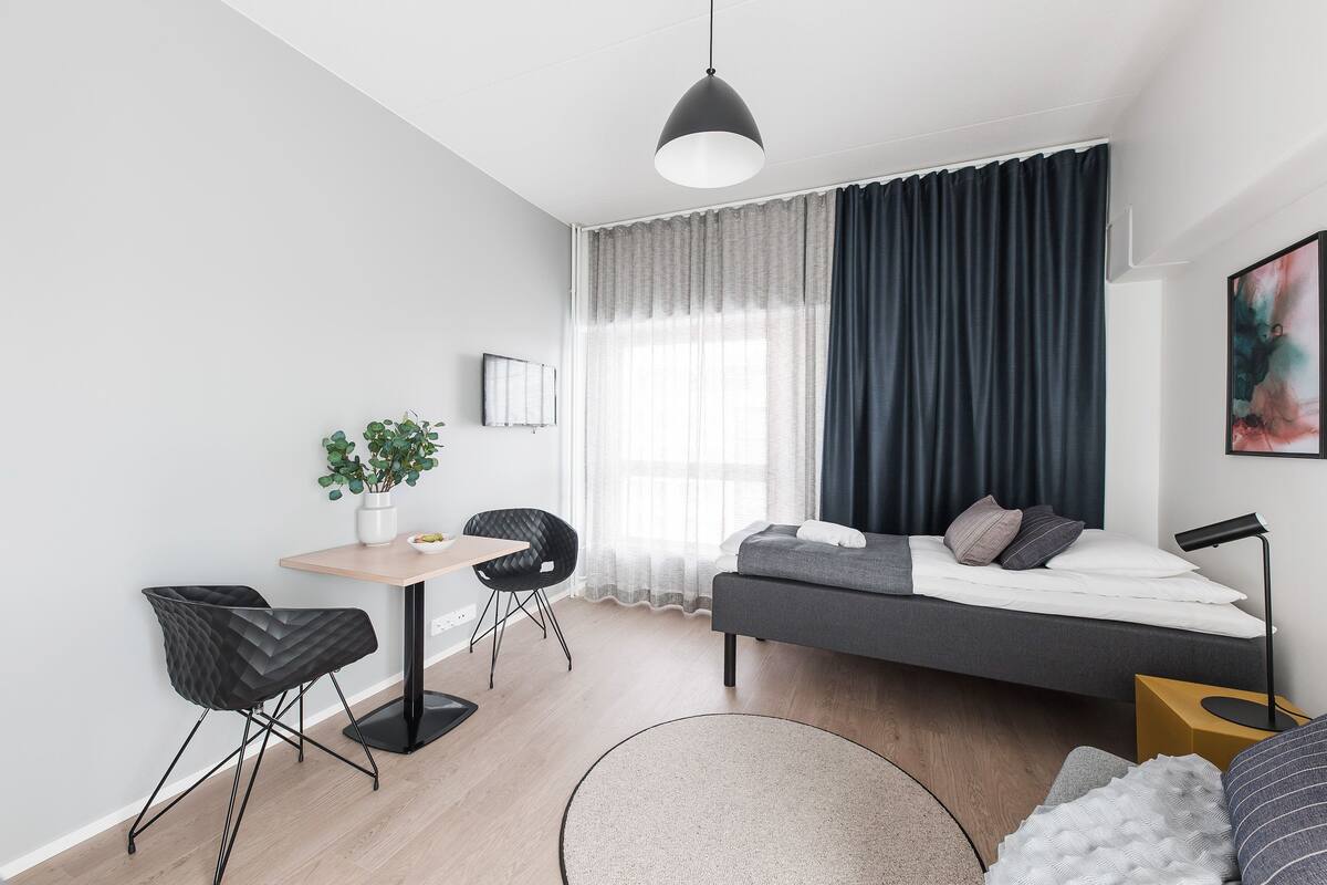 Een moderne, compacte leefruimte met een bed, een klein tafeltje, een stoel, een bank en een potplant.