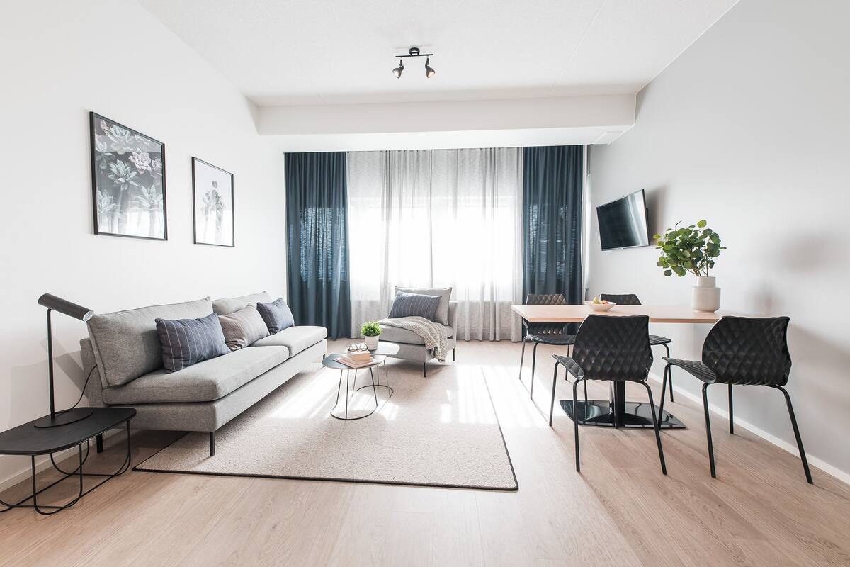 Een moderne woonkamer met een bank, salontafel en stoelen, een eethoek met een tafel en stoelen, een aan de muur bevestigde televisie en een groot raam met gordijnen.