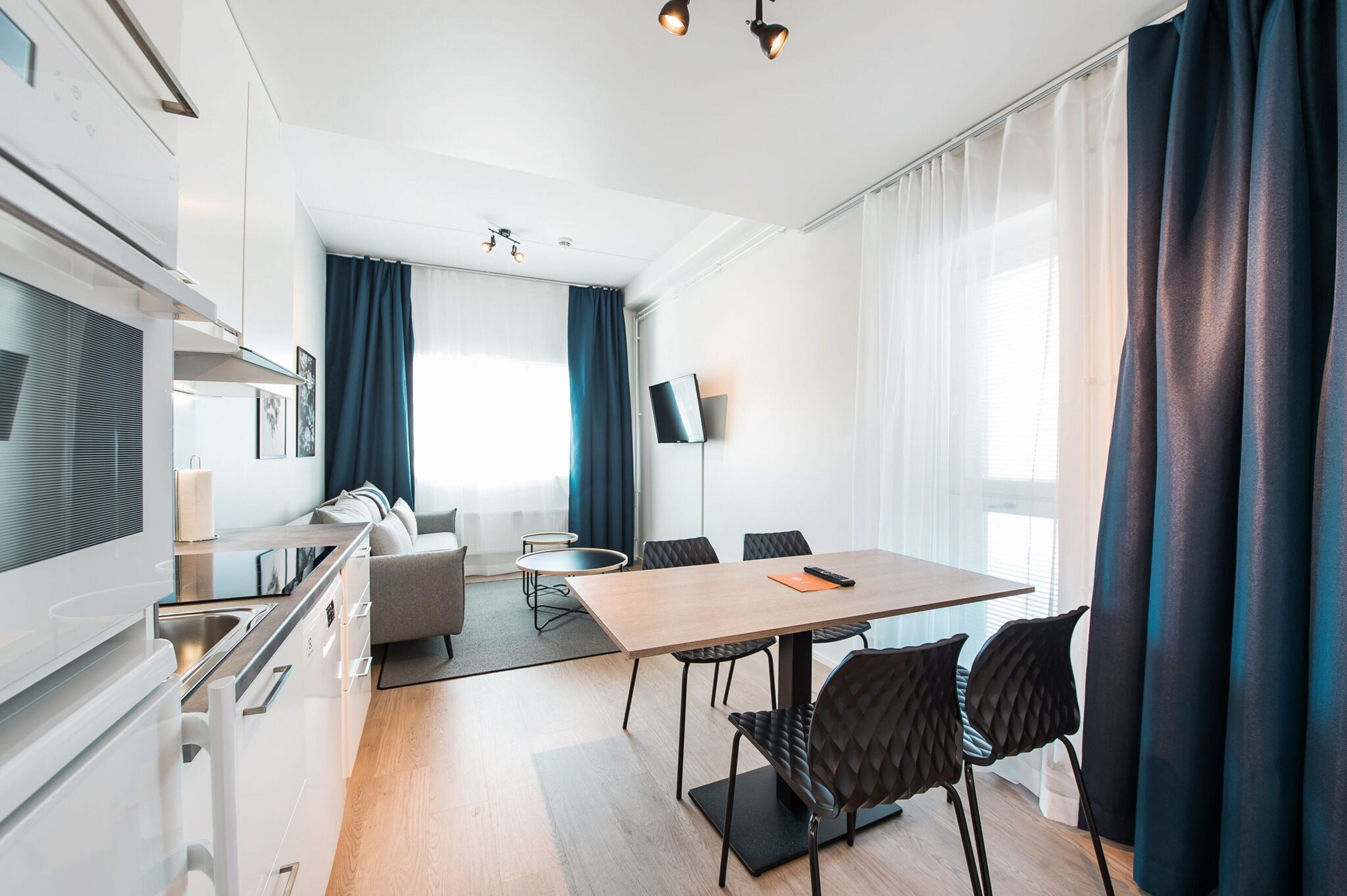 A modern hotel room with a dining area, kitchenette, and a large window with curtains.
