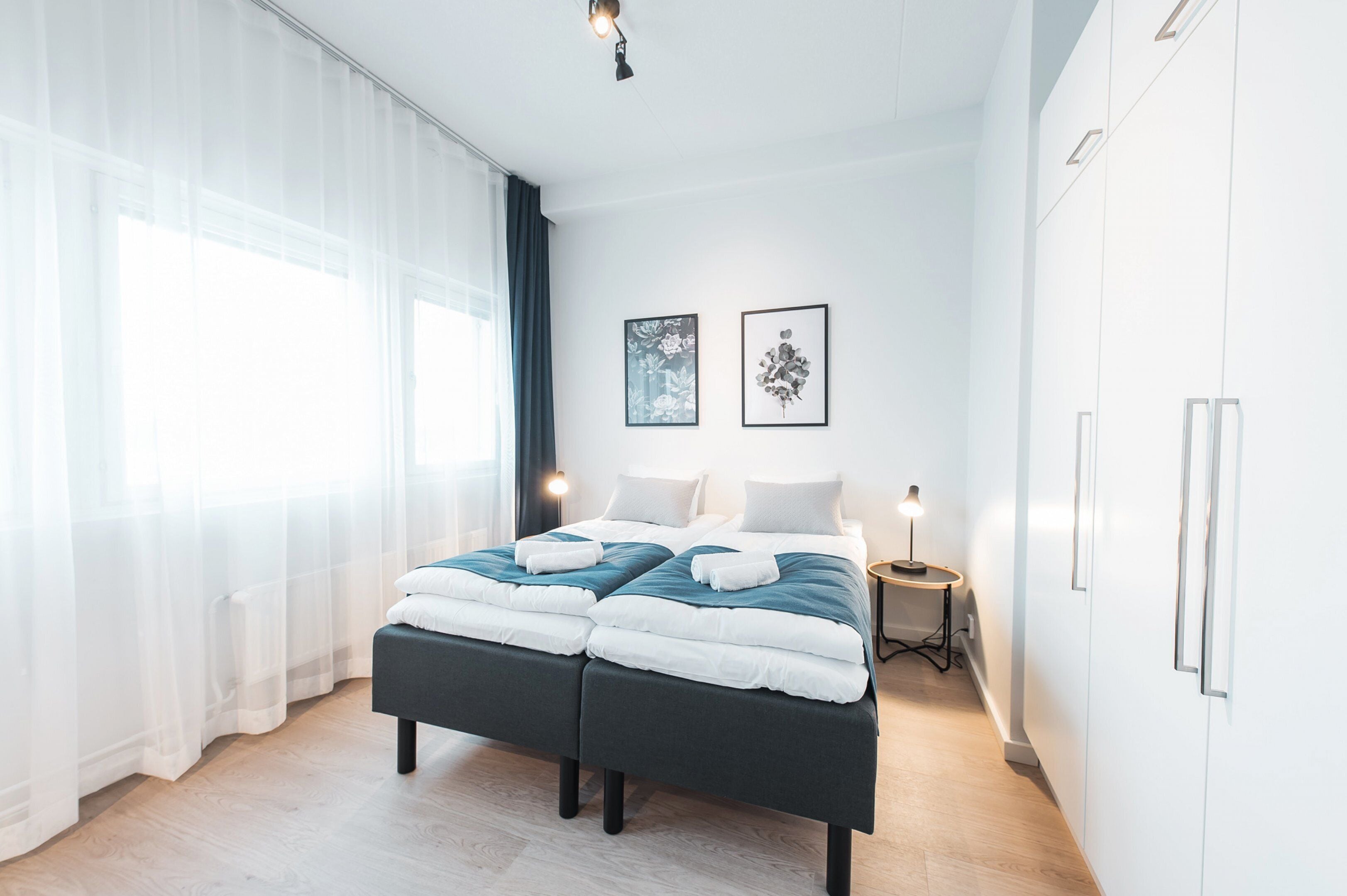 A modern bedroom with a double bed, a bedside table, a lamp, and two framed floral artworks on the wall.