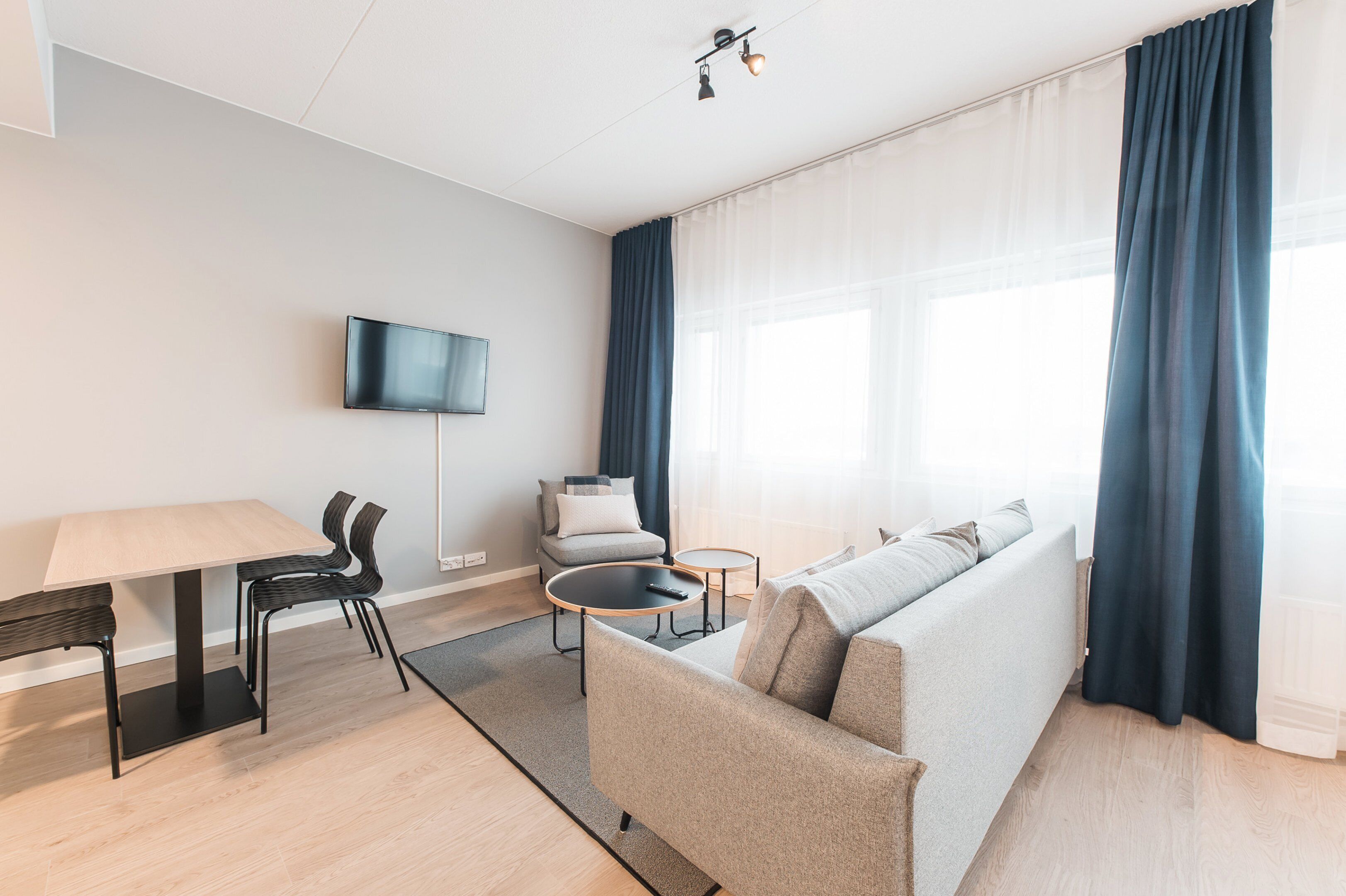Un salon moderne comprenant un canapé, une table à manger avec des chaises, un téléviseur à écran plat fixé au mur et une grande fenêtre avec des rideaux.