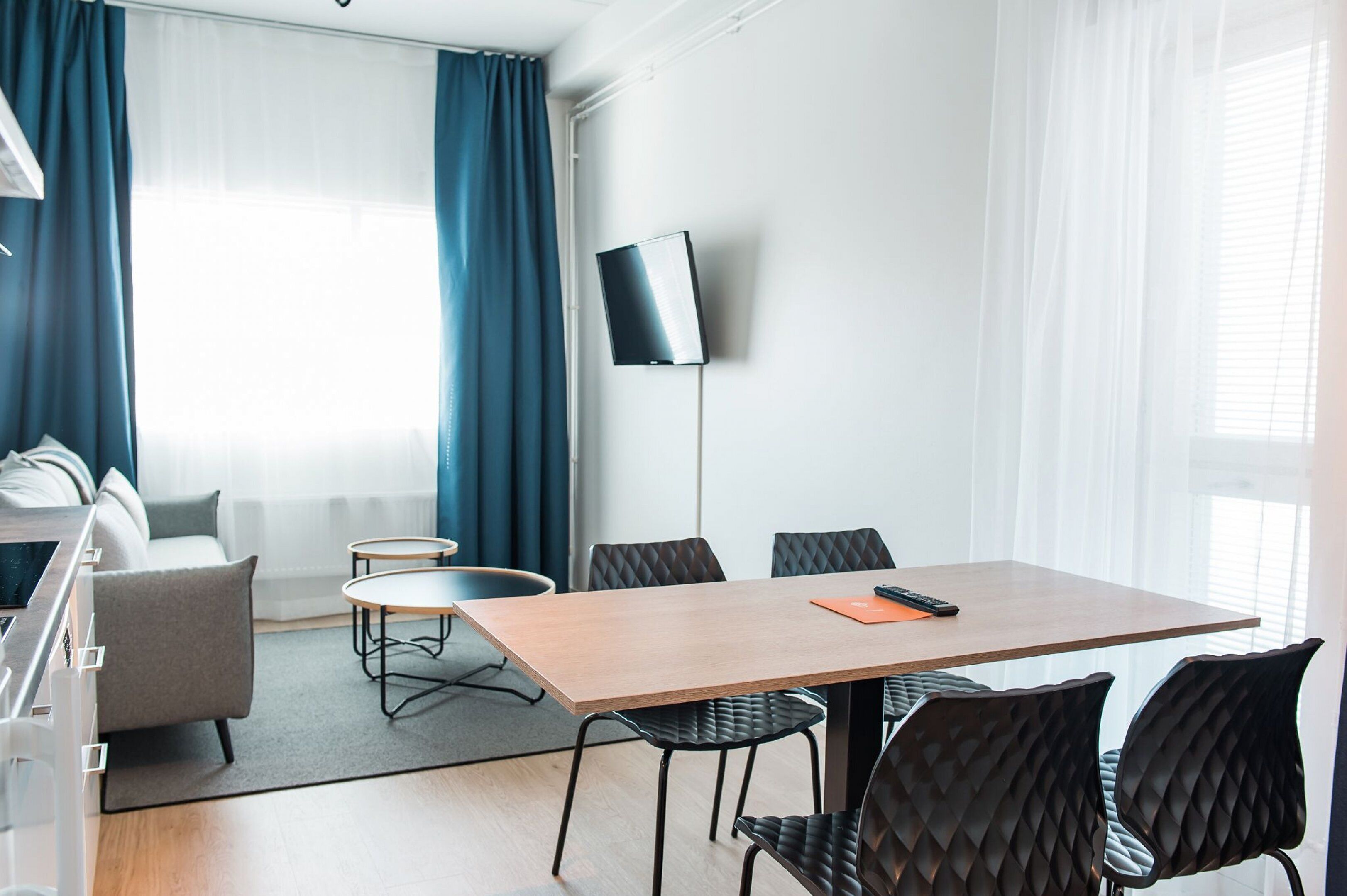 A modern hotel room with a wooden dining table, black cushioned chairs, a flat-screen TV mounted on the wall, and a sofa in the background.