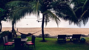 Un espacio de descanso frente a la playa con mesas, sillas y una hamaca, rodeado de palmeras.