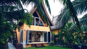 Una casa moderna de dos pisos con balcón, rodeada de abundante follaje y palmeras.
