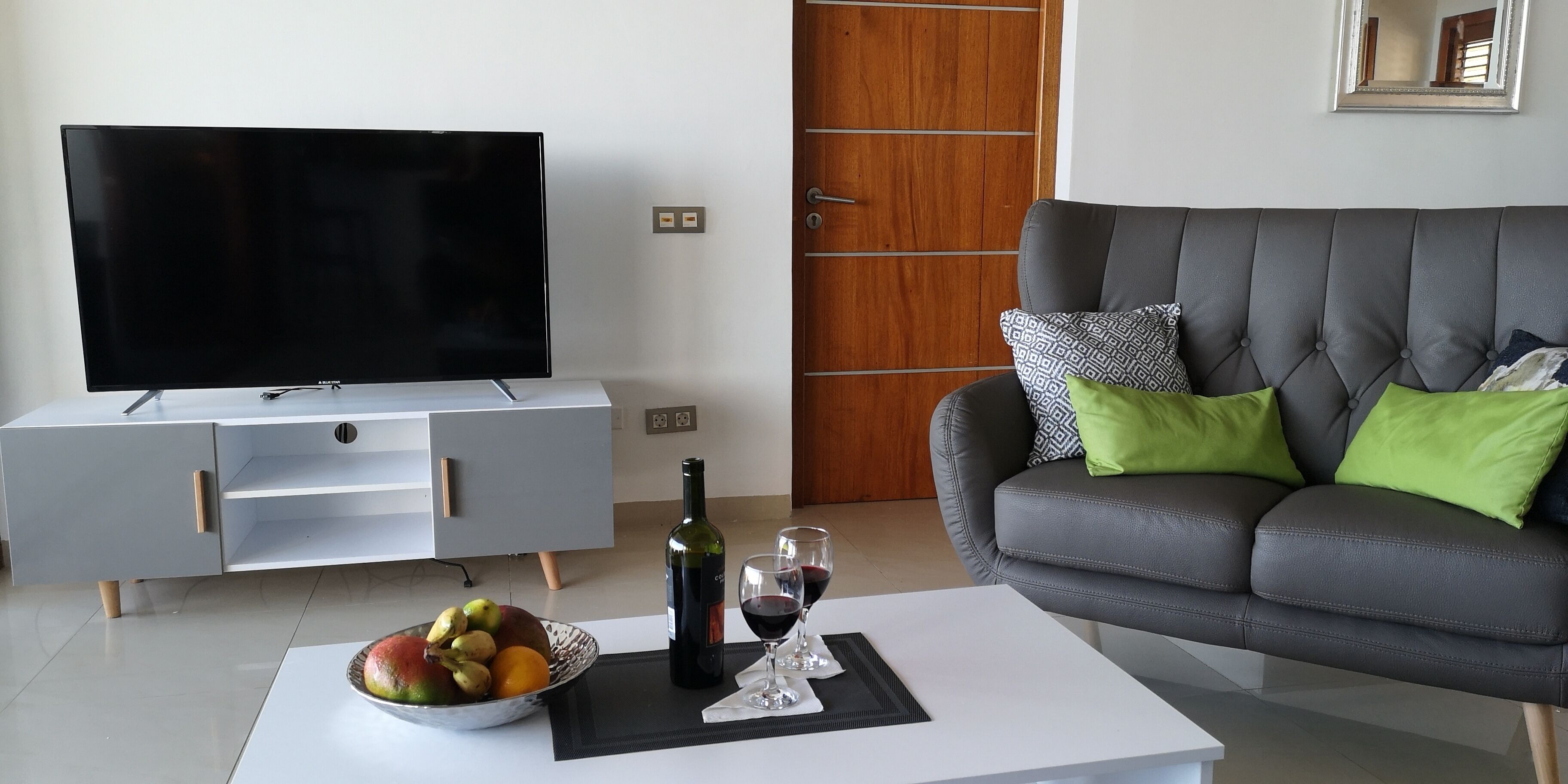 Una sala de estar moderna con un televisor de pantalla plana, un sofá gris con cojines verdes, una mesa de centro con una botella de vino y una copa, y un frutero.