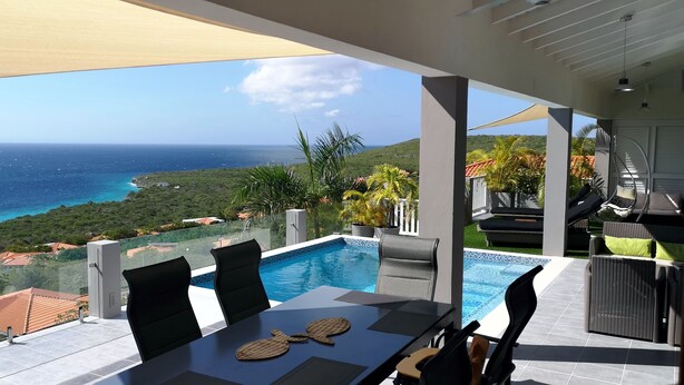 Zona junto a la piscina con vista al océano, una mesa con sillas y un área de estar cubierta.