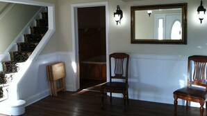 Una habitación con escalera de madera, una silla de madera, un radiador blanco y un espejo en la pared.