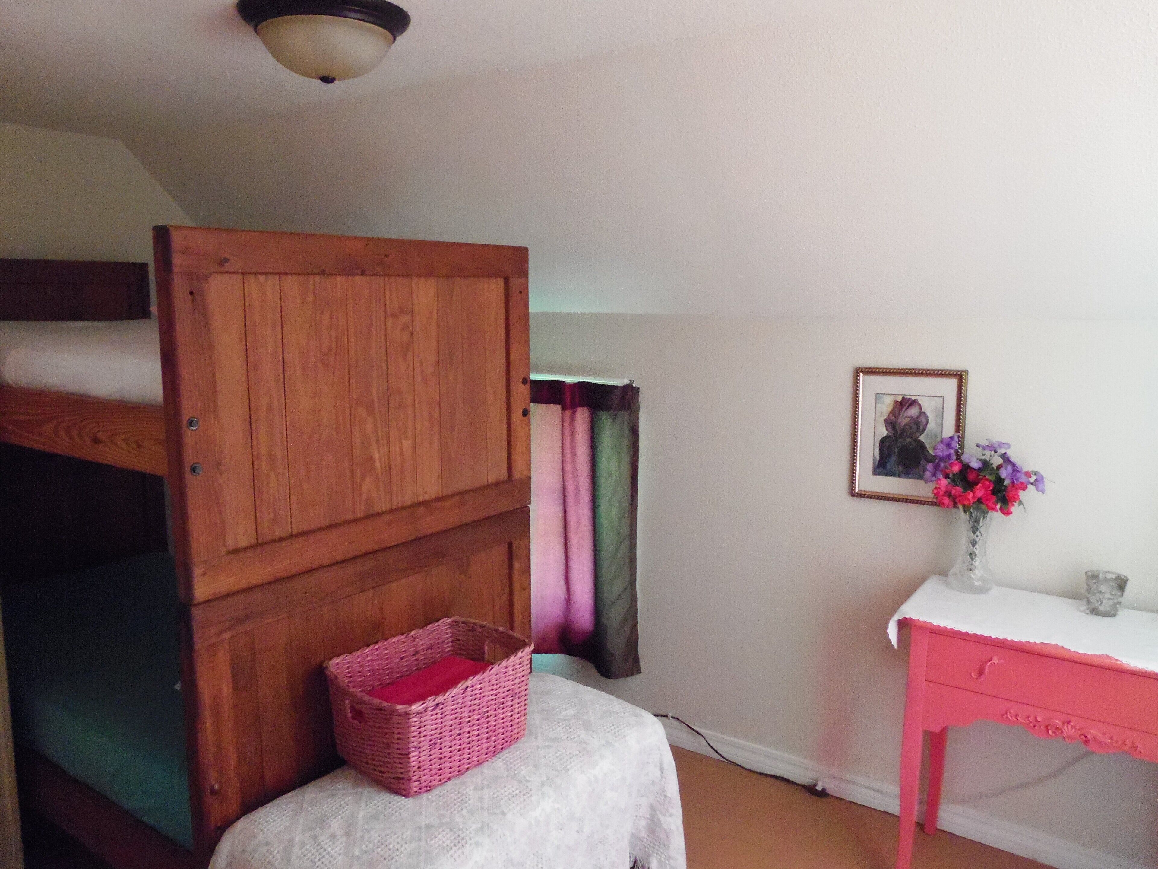 A room with a wooden wardrobe, a small pink basket, a white table with a vase of flowers, and a framed picture on the wall.