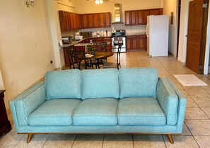 Uma sala de estar com sofá azul claro, armários de madeira, cozinha com fogão e geladeira, e uma mesa de jantar com cadeiras.