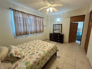Quarto com cama, penteadeira, ventoinha no teto e porta que dá para outro cômodo.