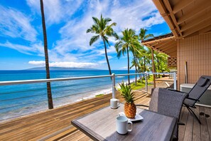 A wooden deck with a table and chairs overlooking a beach with palm trees and the ocean. - Charming 3 BR Puamana Oceanfront in private gated community (Lahaina)
