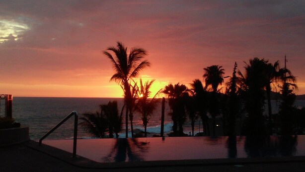A sunset view over a pool area with palm trees and a distant ocean. -  Stunning ocean and one of Mazatlans top tourist attractions cliff divers view. (Mazatlán)