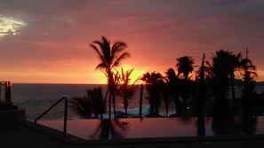 A sunset view over a pool area with palm trees and a distant ocean.