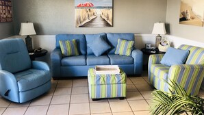 A living room with a blue sofa, striped ottoman, and matching armchair, a ceiling fan, and a beach-themed painting on the wall. - #404- 2 bed-1 bath sleeps 6- Fall beach getaway…& Winter Snowbirds! Book now! (Gulf Shores)