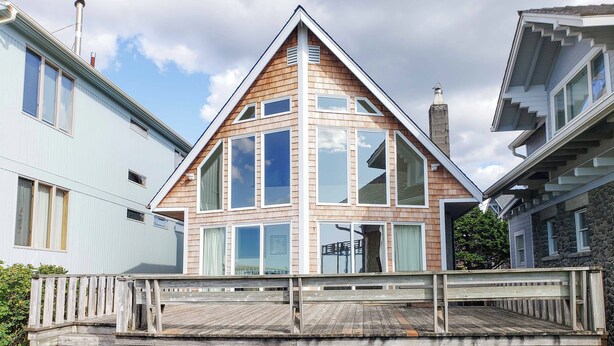 Une maison moderne dotée d’un toit triangulaire imposant, de grandes fenêtres et d’une terrasse en bois à l’avant.