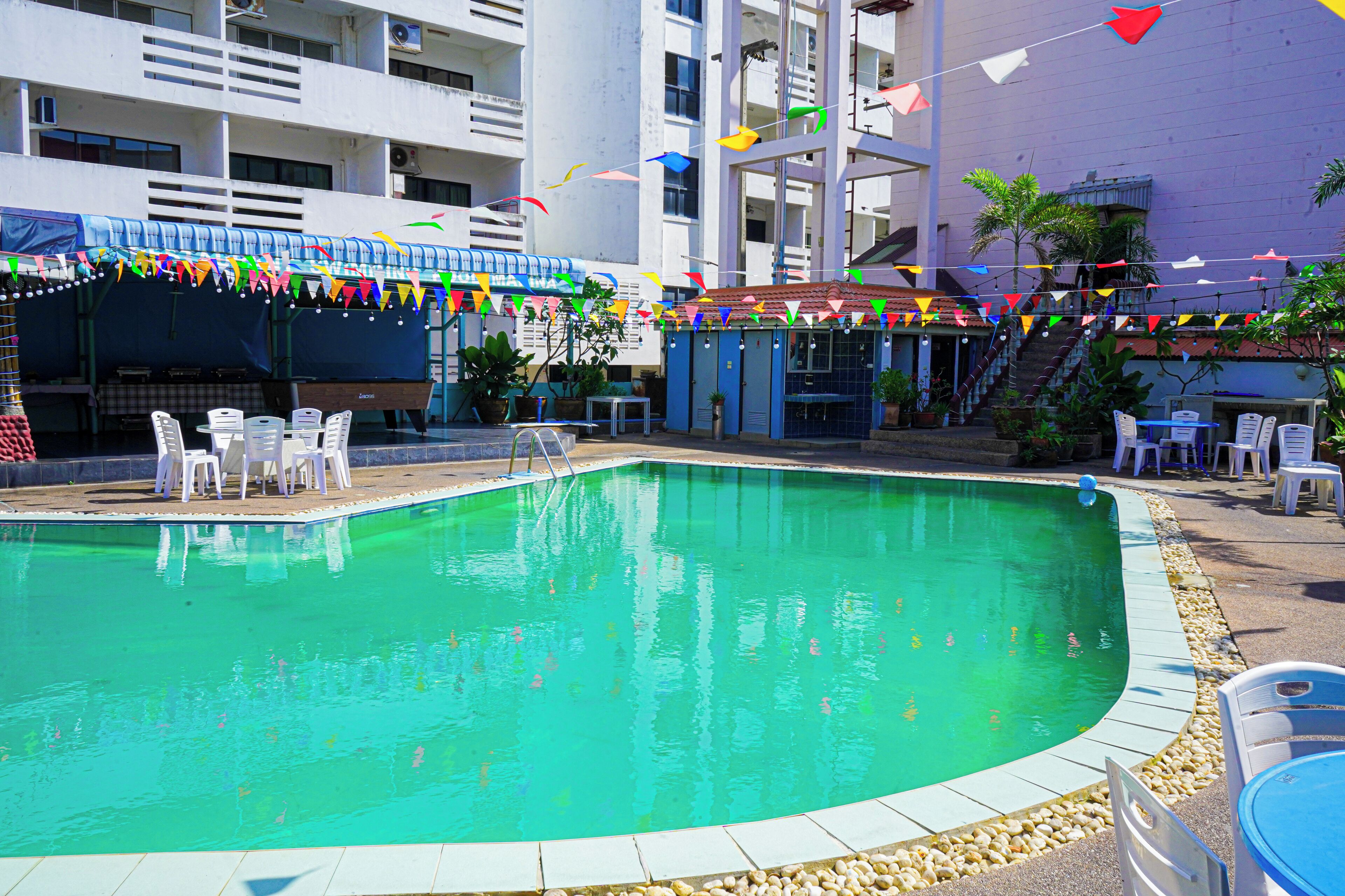 A swimming pool with a tiled edge, surrounded by white plastic chairs and tables, and a building with balconies in the background.