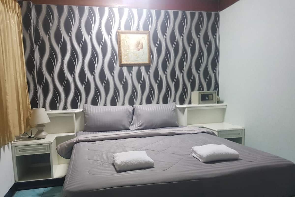 A bedroom with a patterned wallpaper, a bed with pillows, a bedside table with a lamp, and a framed picture on the wall.