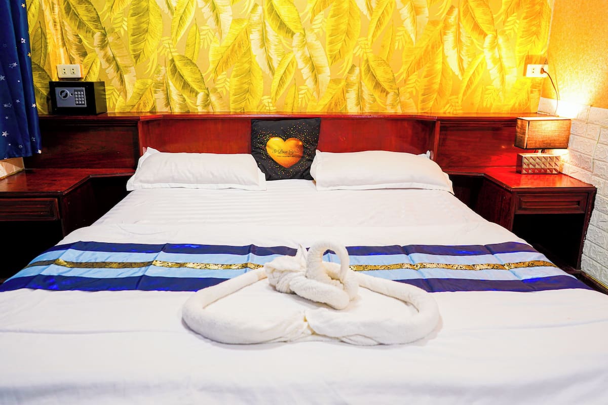 A hotel room with a large bed, a heart-shaped pillow, a swan-shaped towel, and a wall with a banana leaf pattern.
