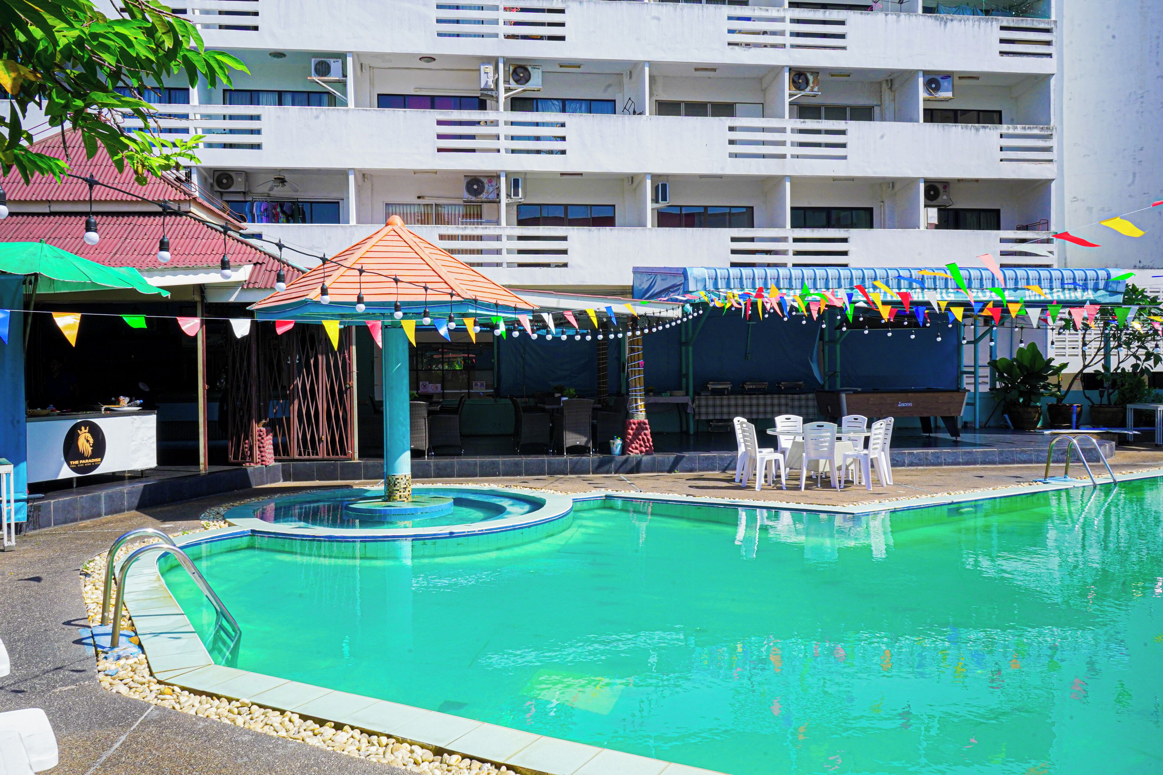 A swimming pool with a tiled edge, surrounded by white lounge chairs and a covered area with a red roof.