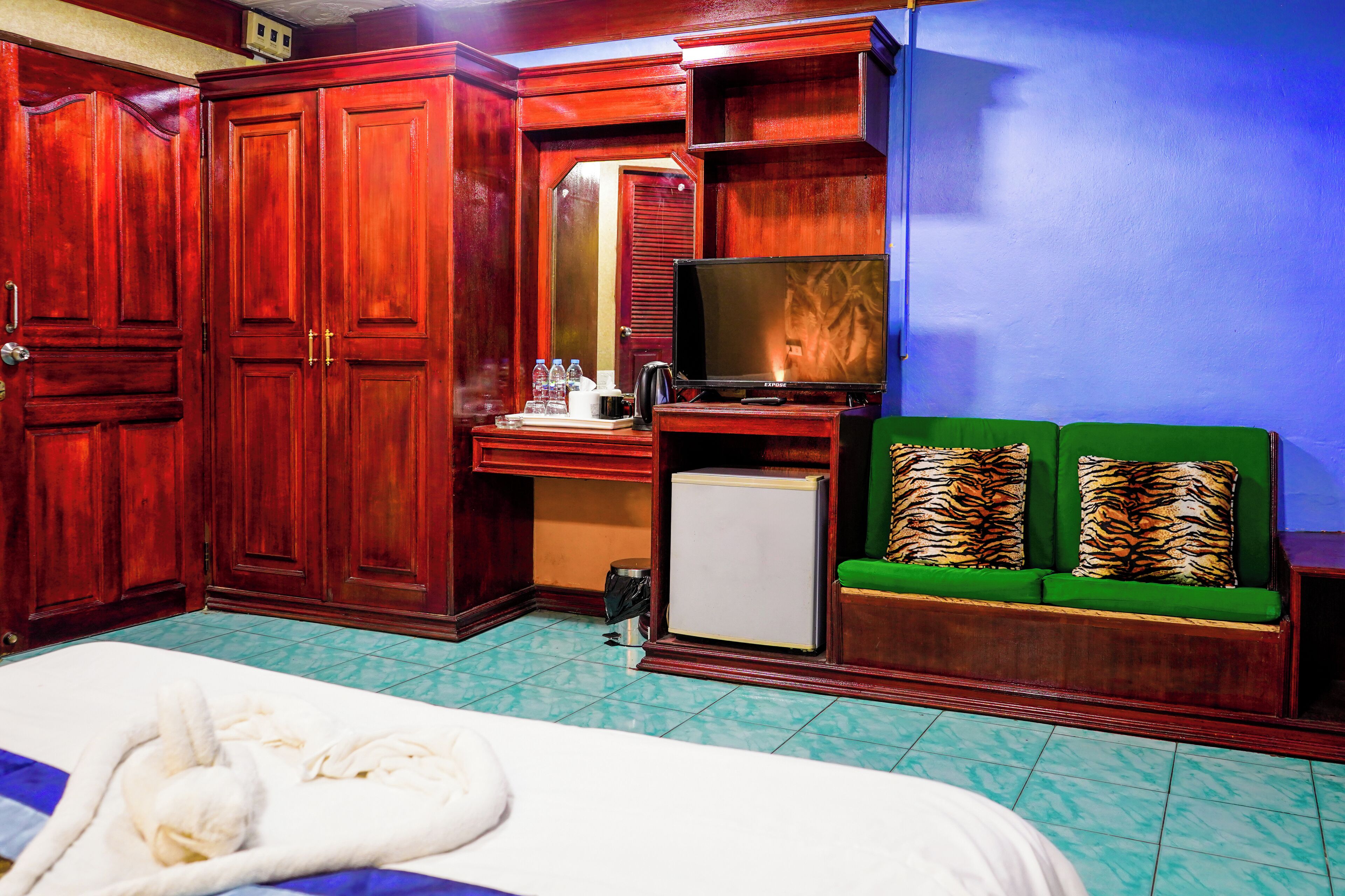 A hotel room with a bed, a desk with a television, a small refrigerator, and a green sofa with tiger-patterned cushions.