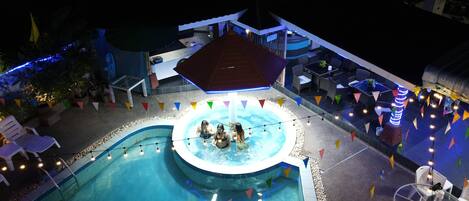 An outdoor swimming pool area with a hot tub, surrounded by lounge chairs and tables, and decorated with colorful flags.