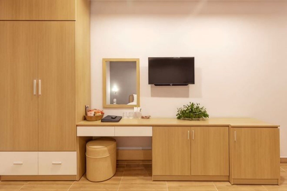 A room with a wooden wardrobe, a desk with a mirror, a television mounted on the wall, and a trash bin.