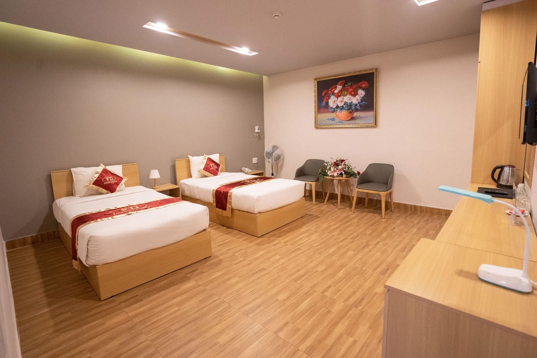 A hotel room with two beds, a desk, a chair, a small table with flowers, and a painting on the wall.