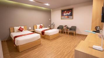 A hotel room with two beds, a desk, a chair, a small table with flowers, and a painting on the wall.