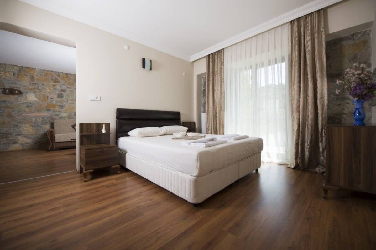 A bedroom with a large bed, wooden flooring, a stone wall, and a view of the outdoors.