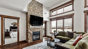 A living room with a stone fireplace, a large TV, a green sofa, and a view of the mountains through large windows. - Top-Floor Luxury Condo with Panoramic Views at Trailhead Lodge! (Steamboat Springs)