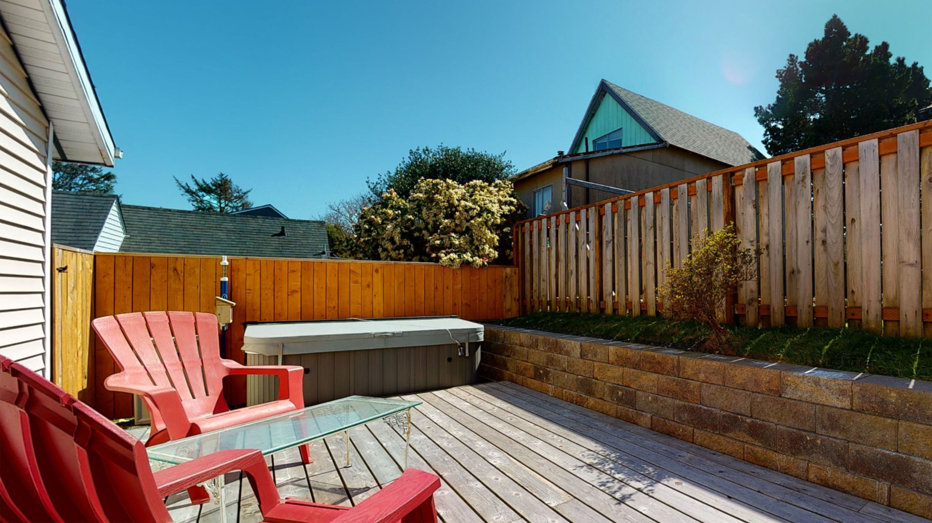 A backyard patio with a glass table, red chairs, a hot tub, and a wooden fence.