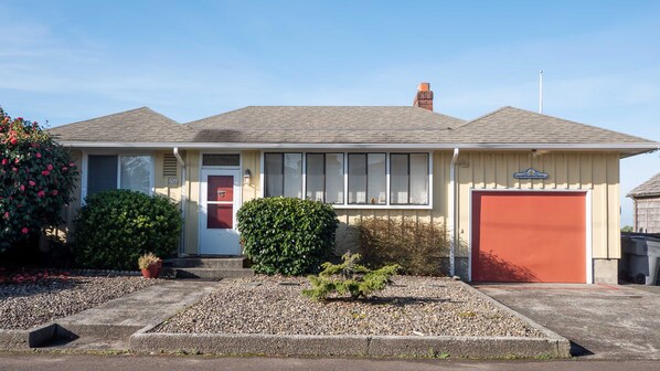 A single-story house with a red garage door, a front porch with steps, and a small front yard with gravel and shrubs.